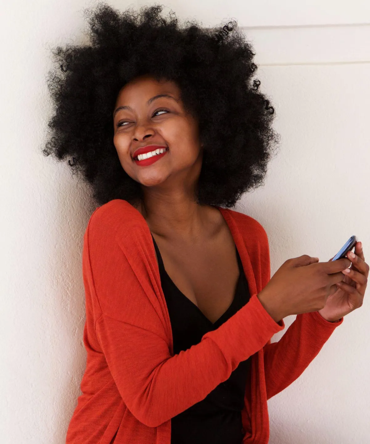 Smiling woman with curly hair wearing a red cardigan, holding a smartphone and looking relaxed. Represents confidence and comfort after breast reduction surgery.
