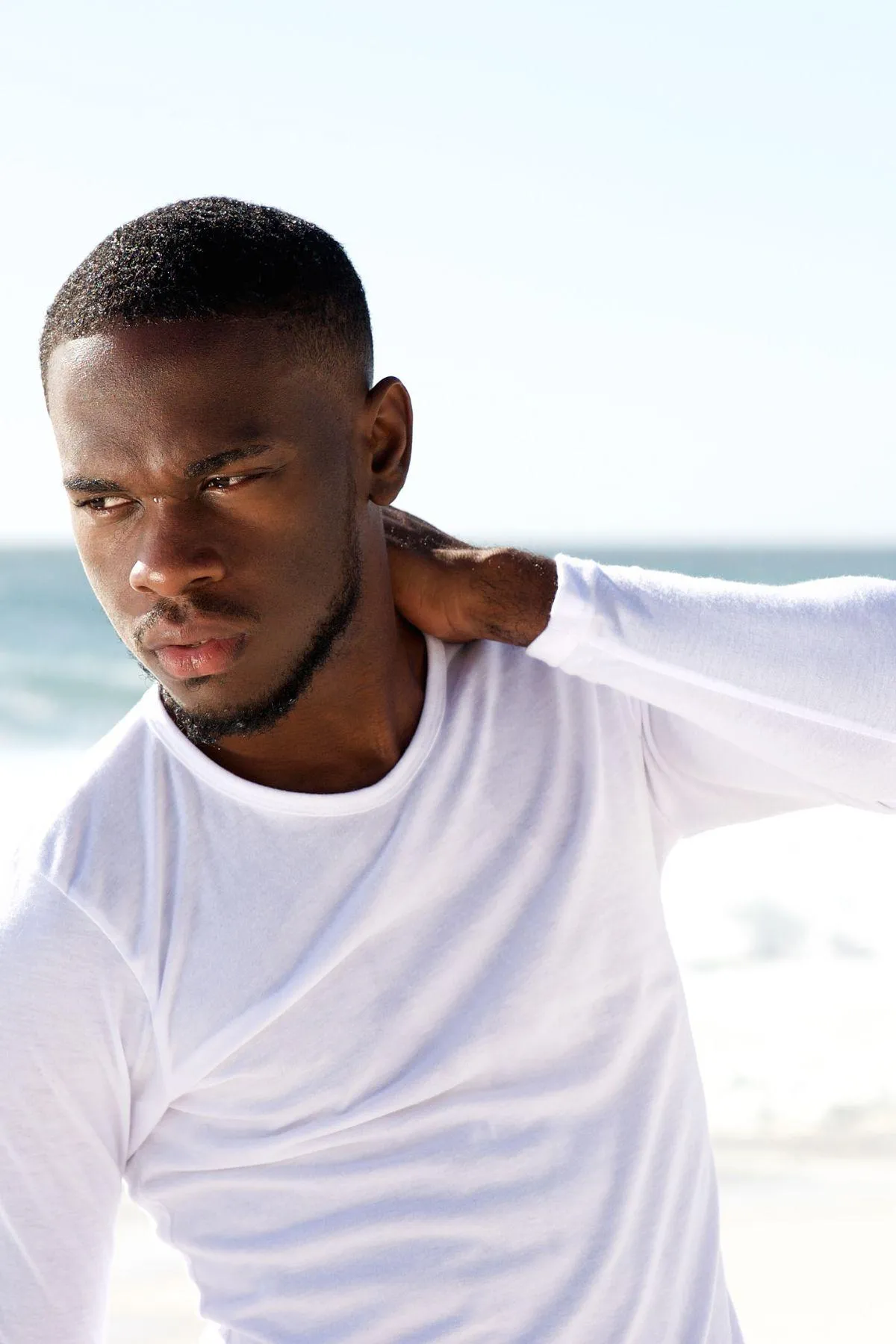 Man standing outdoors near the ocean, wearing a white long-sleeve shirt, with a contemplative expression and hand resting on the back of his neck—highlighting masculine features and body confidence in a serene beach setting.