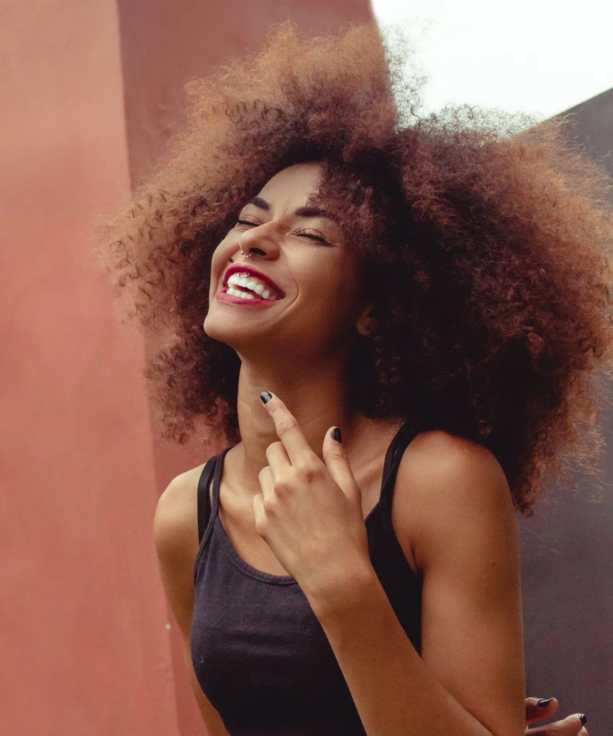 Smiling woman with voluminous curly hair wearing a sleeveless black top, laughing with head tilted back and hand raised near her face—highlighting expressive eyes and natural beauty associated with blepharoplasty results.