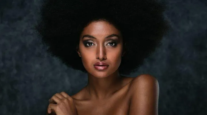 Woman with voluminous afro hairstyle posing elegantly against a dark textured background, with glowing skin and confident gaze—highlighting refined facial contours and poise associated with buccal fat removal results.