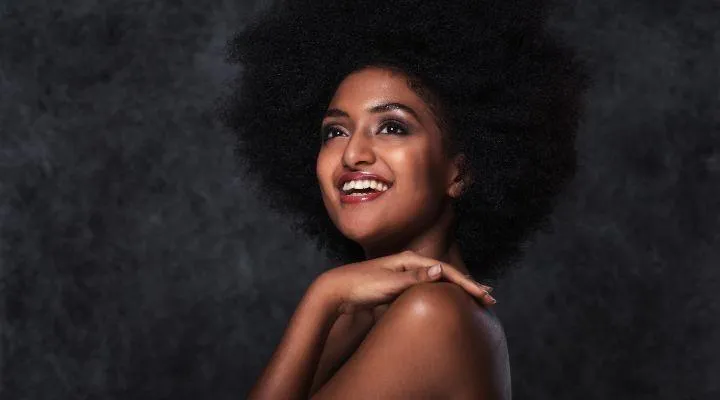 Smiling woman with voluminous natural curls wearing a sleeveless black top, gently resting her hands on her shoulder against a dark background—highlighting facial symmetry and confidence, ideal for showcasing ear lobe repair results.