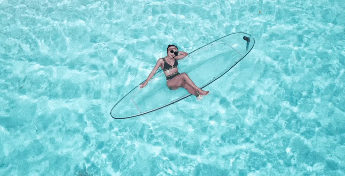 Woman lying on a transparent kayak over crystal-clear turquoise water, wearing a black and white swimsuit and sunglasses, appearing relaxed and serene in a tropical setting.