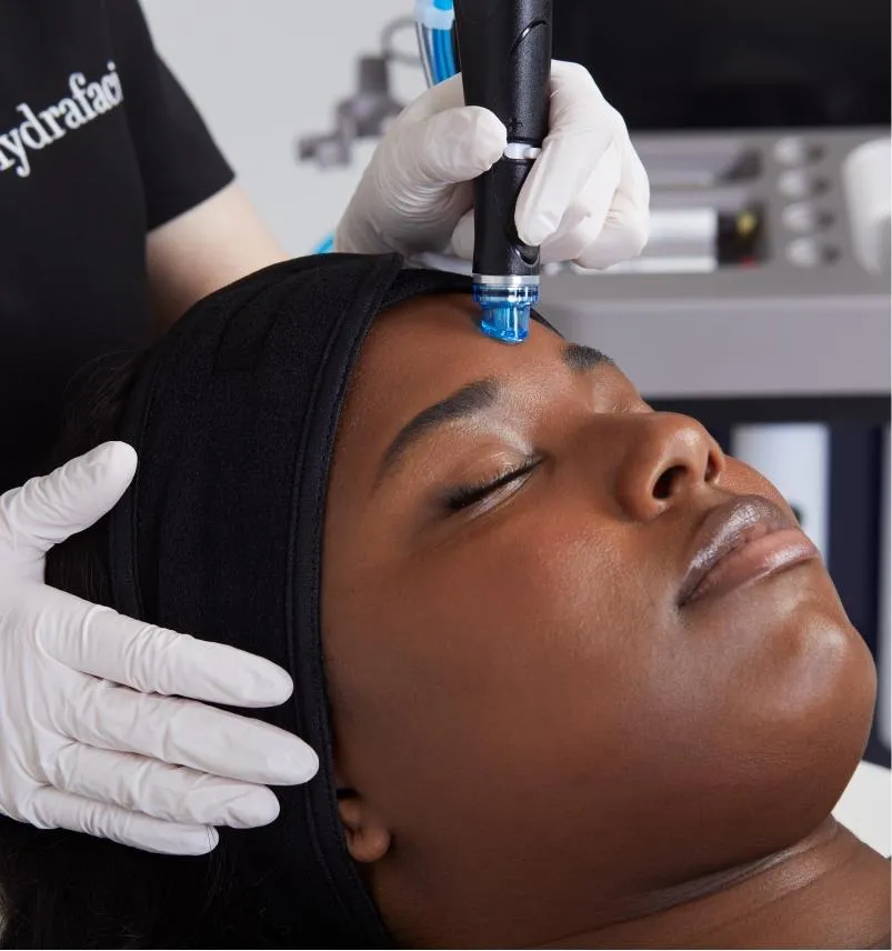 Alt text suggestion: Close-up of a HydraFacial treatment being performed on a woman's forehead by a professional wearing gloves, using a specialized wand while the patient lies relaxed with eyes closed—highlighting advanced skincare technology and rejuvenation.