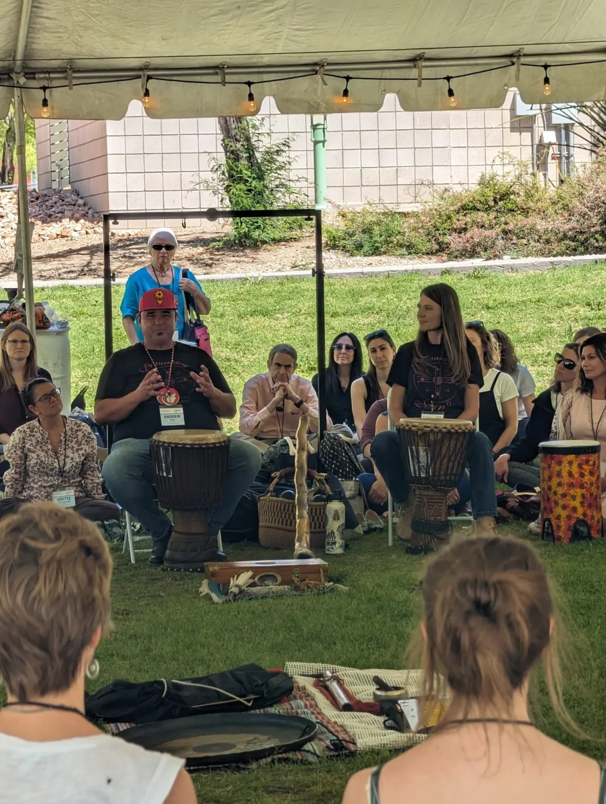 A man from behind leading a diverse group of people sitting in a circle in an industrial loft space with exposed brick, all playing hand drums.
