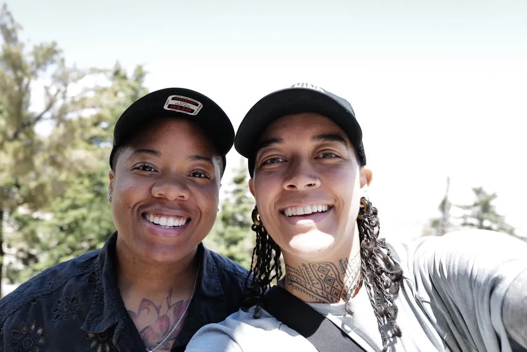 Two masculine presenting women, smiling together in nature with trees in the background