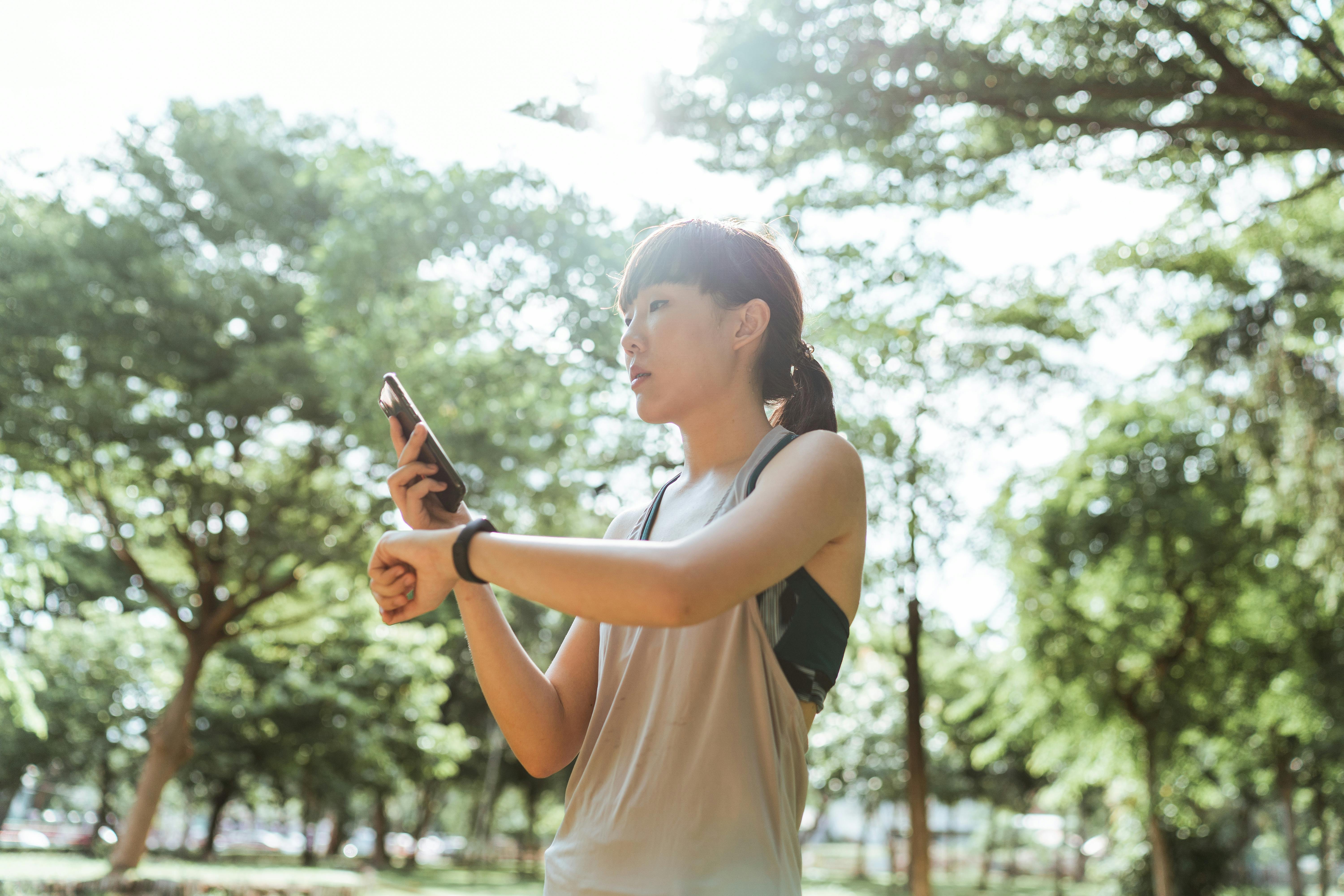 Asian woman in activewear using smartphone and smartwatch outdoors in a sunny park.