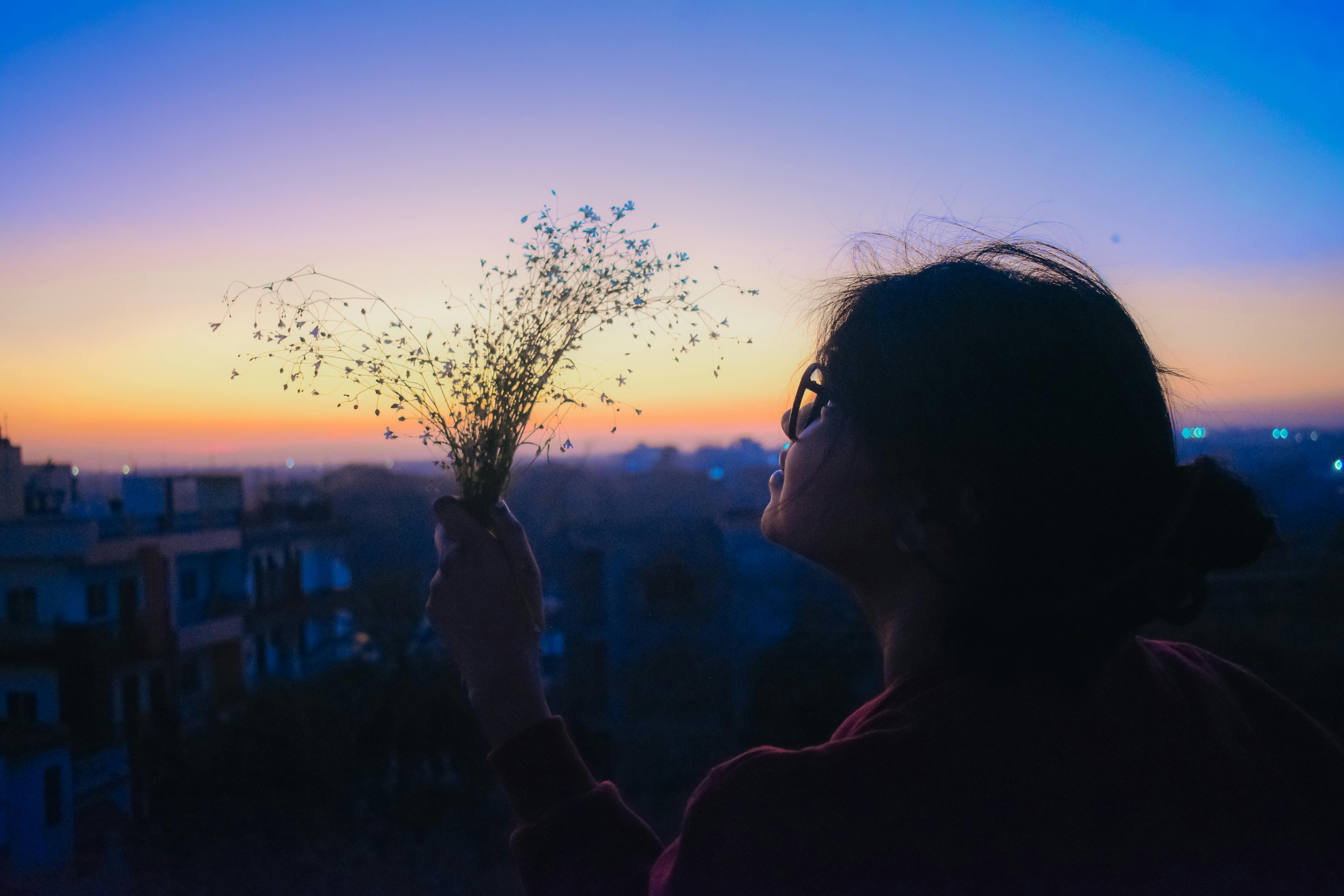 A woman holds a bouquet silhouetted against a vibrant sunset in an urban setting.