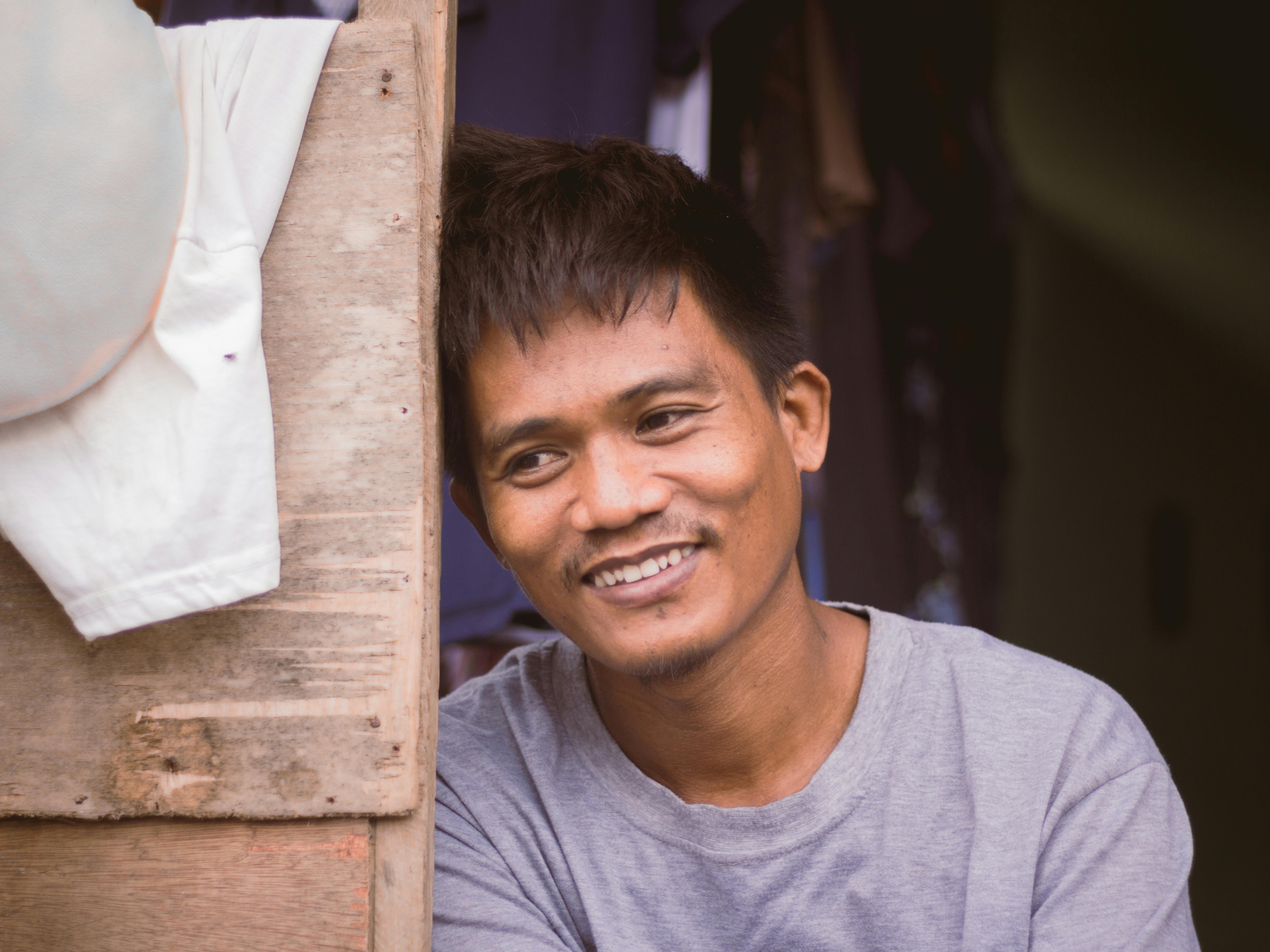 A cheerful Asian man in casual clothing smiles while leaning against a wooden wall.