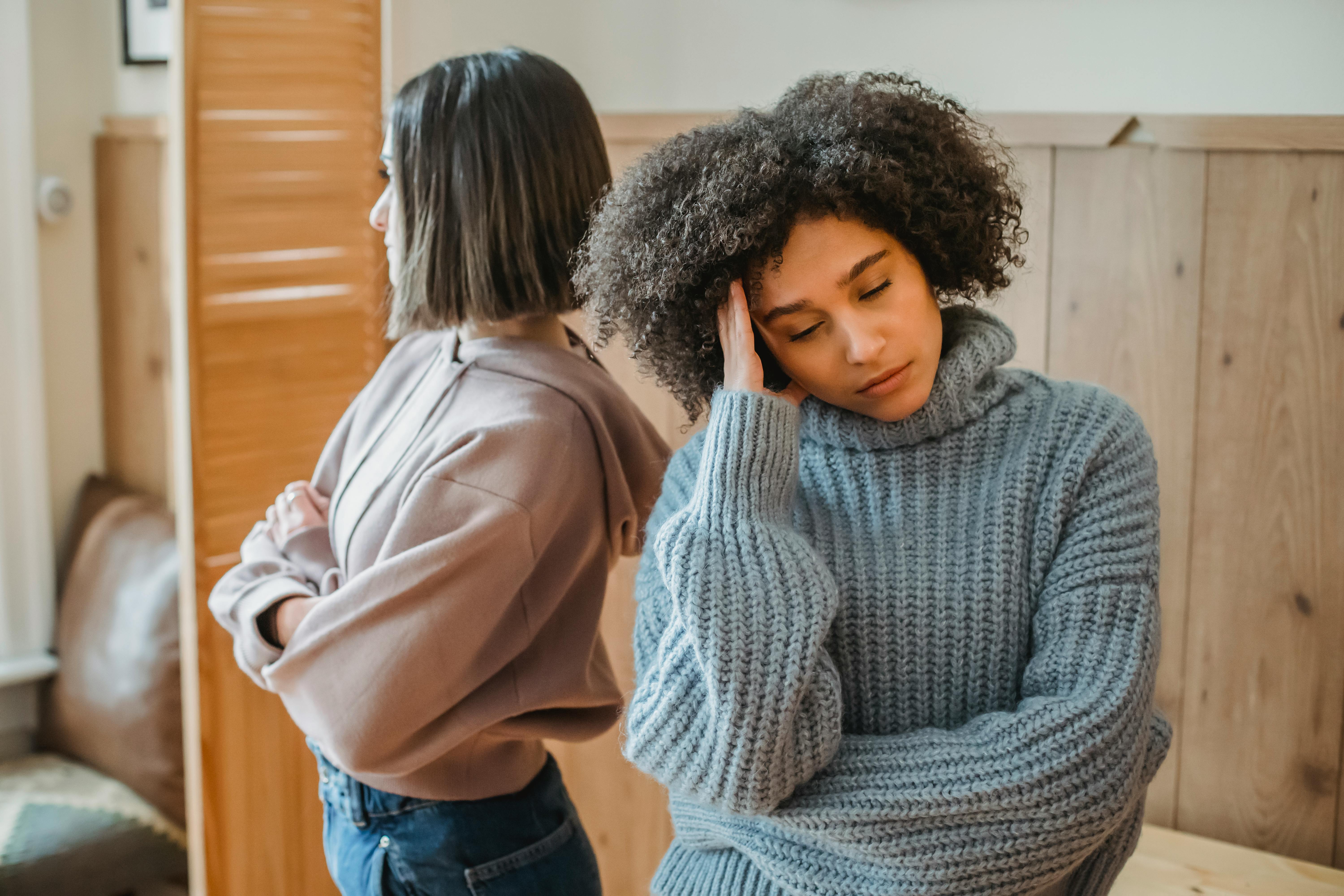 Two women stand back-to-back indoors, exhibiting disagreement and frustration.