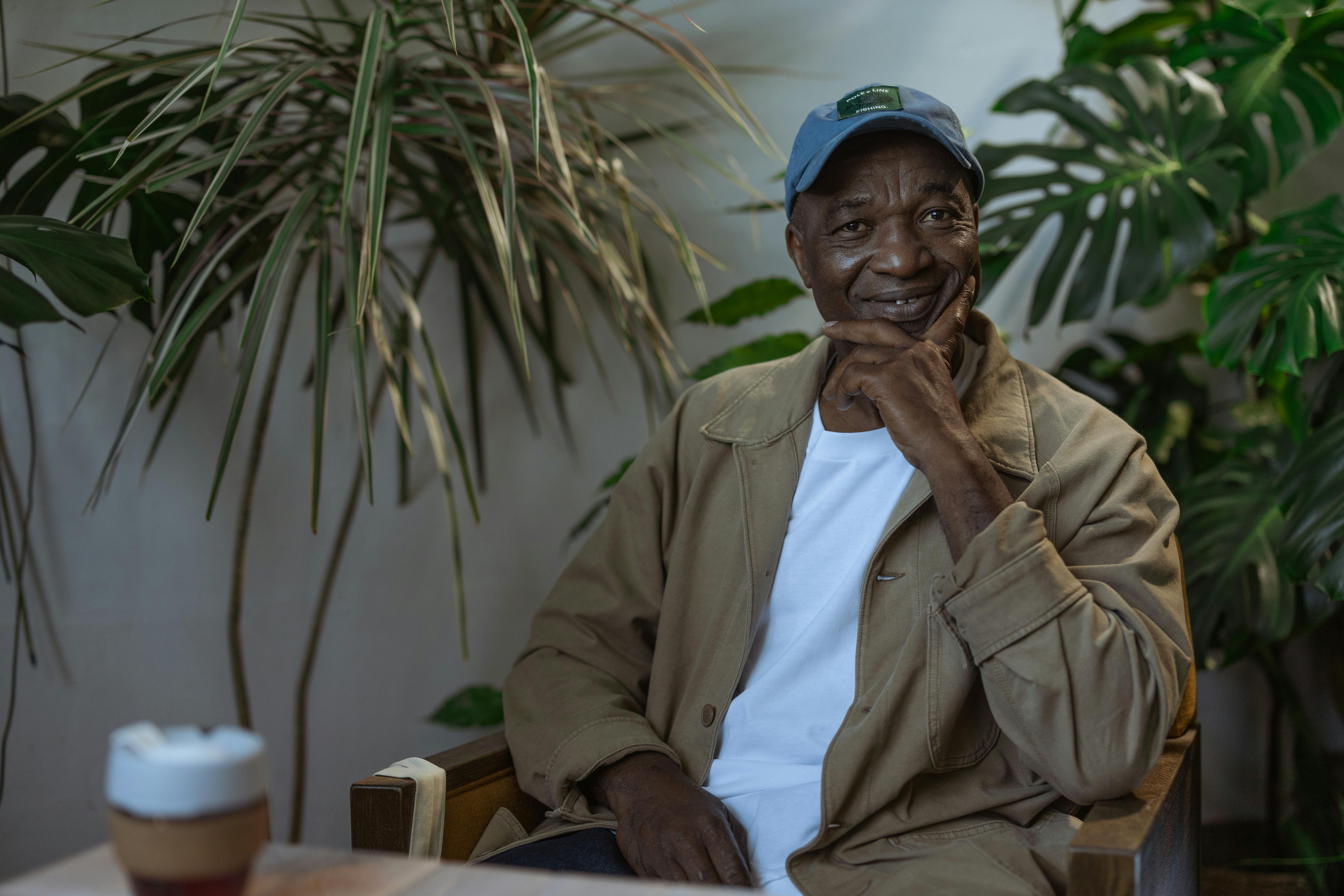 Smiling elderly man in casual attire sitting indoors surrounded by lush plants.