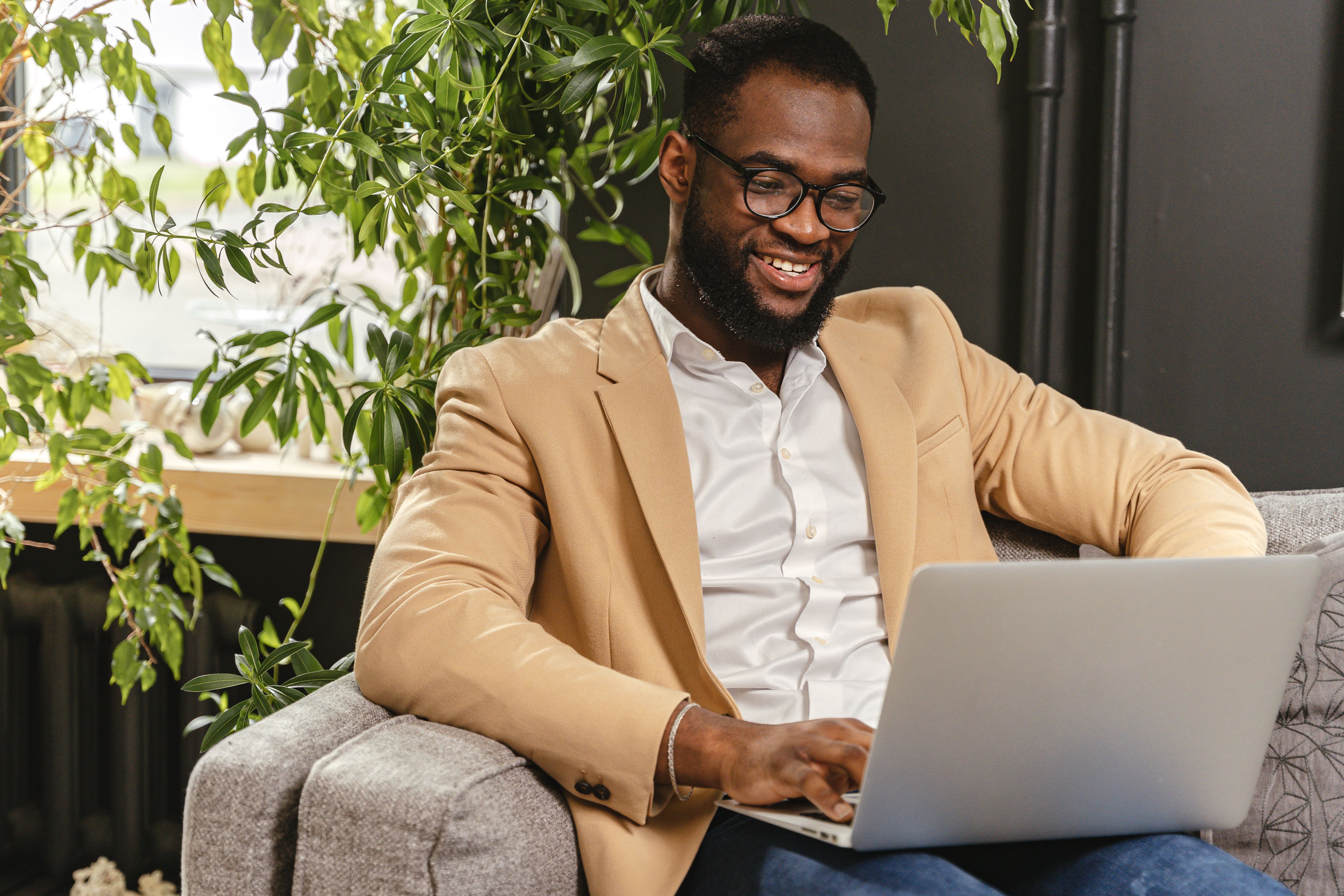 A founder sitting on a couch works remotely on his laptop, smiling in a bright indoor setting.