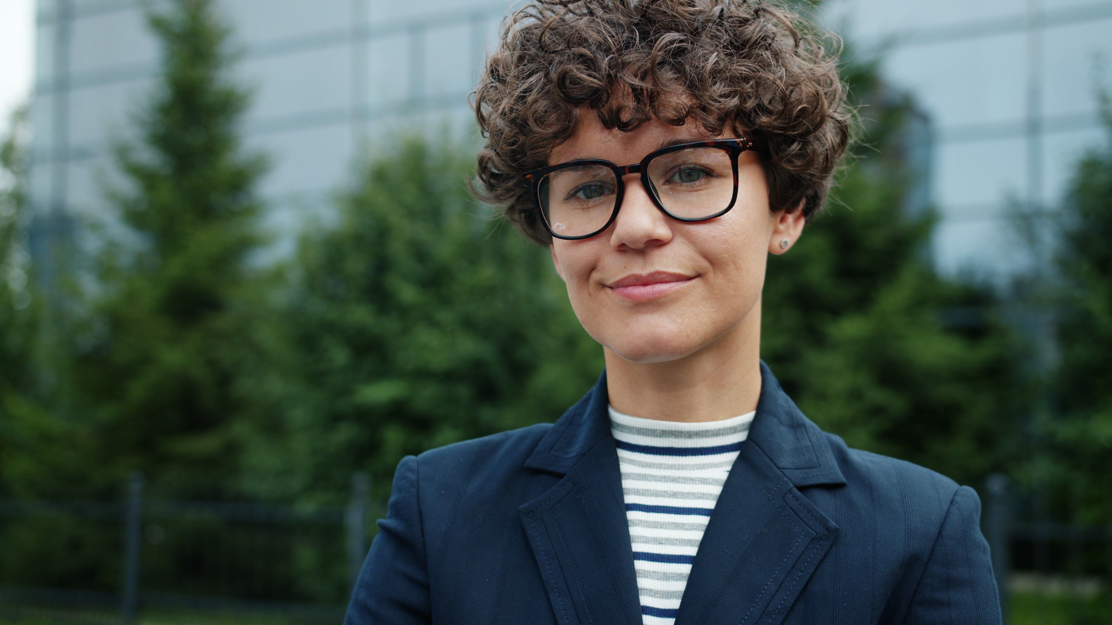 Outdoor portrait of a confident business professional with curly hair and glasses.