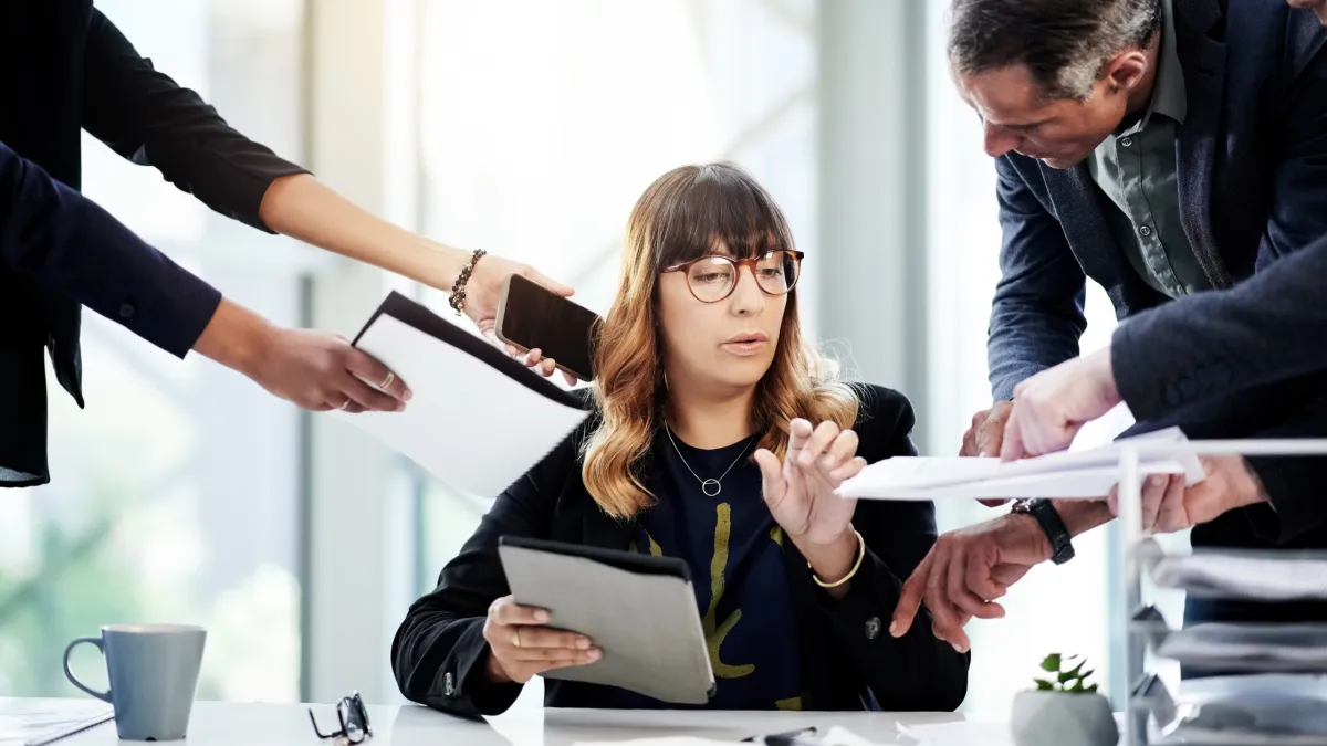 Overwhelmed business professional multitasking at a desk while coworkers hand over documents and devices, representing burnout and the hidden cost of doing everything yourself.