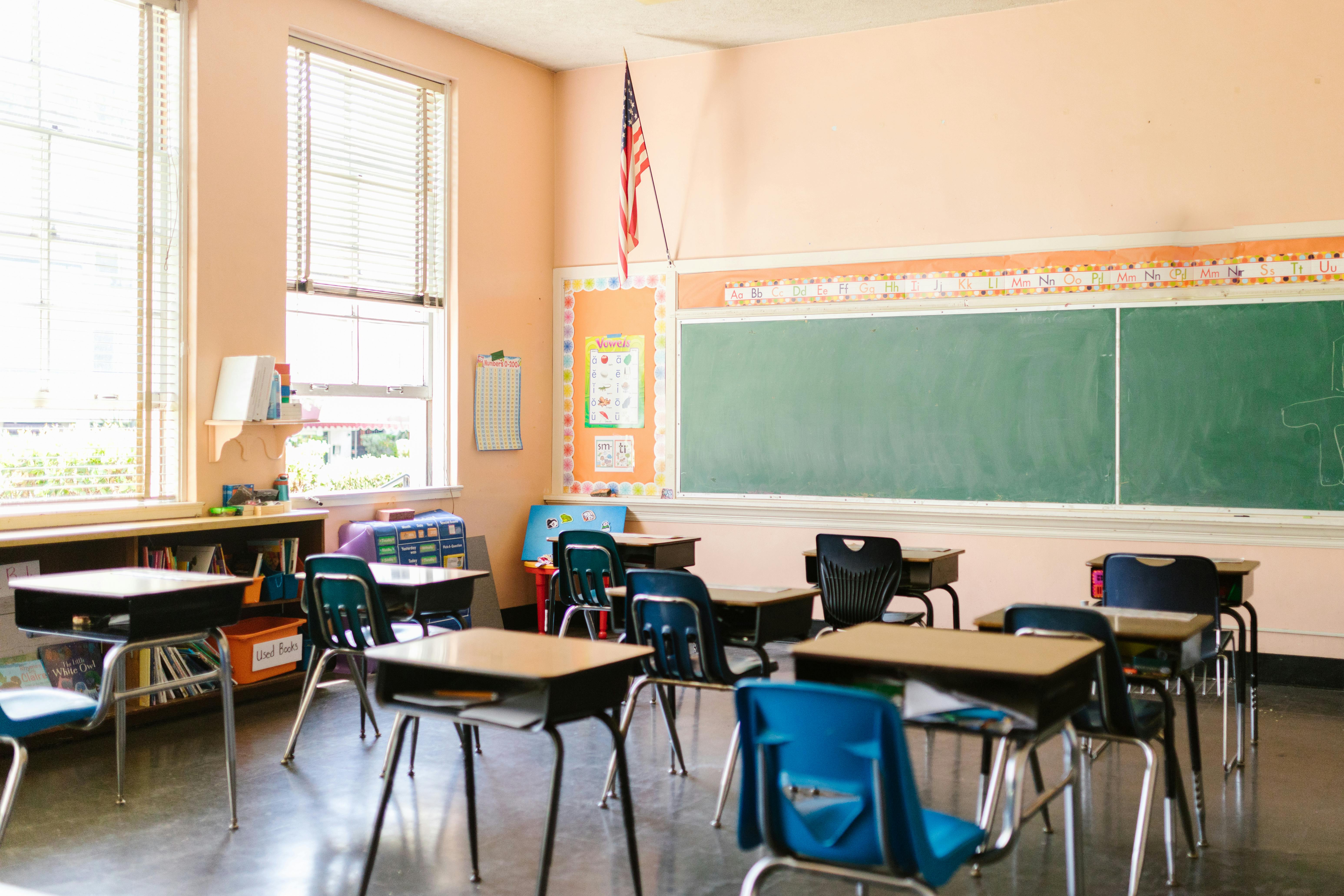 An empty primary school classroom with chairs and tables arranged in rows and a blackboard at the front of the room.