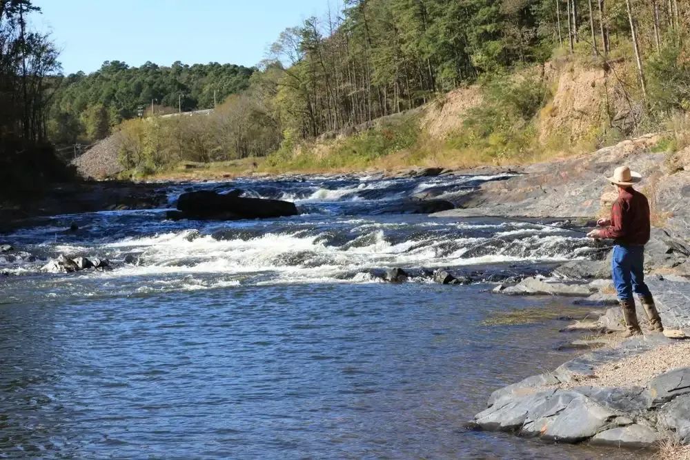 Man fishing on rocky riverbank with forested hills and flowing water.