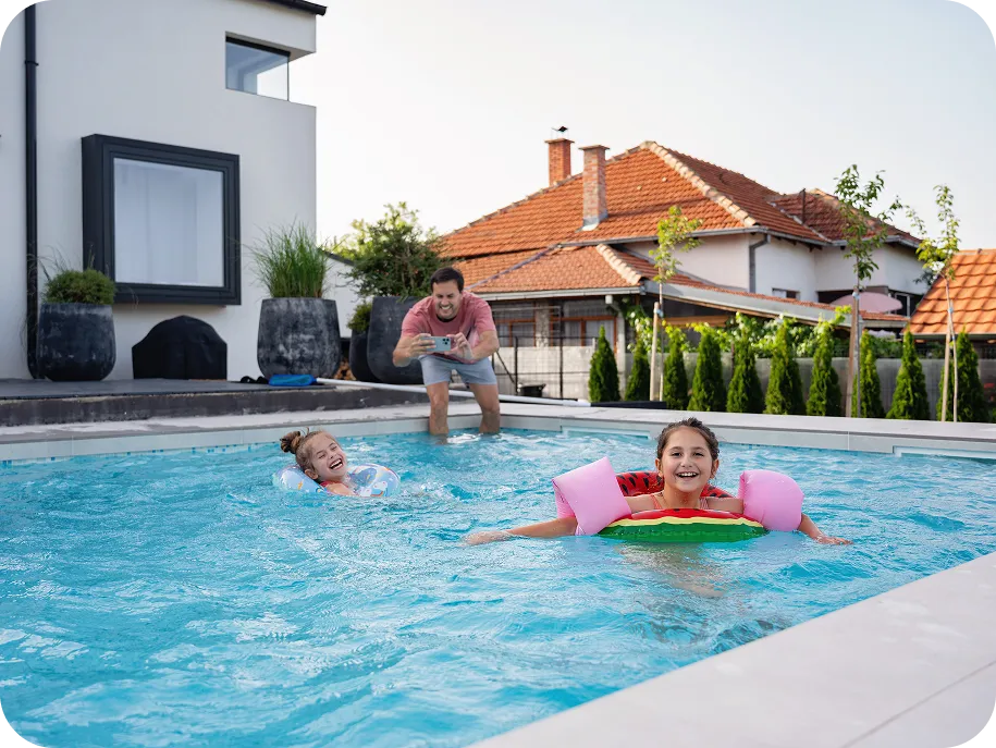 Father taking pictures of his children playing in a backyard pool with inflatable toys.