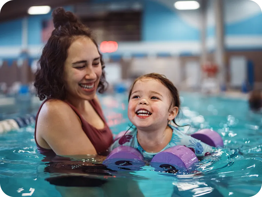 Toddler wearing floaties smiling during a swim lesson with mom in an indoor pool.