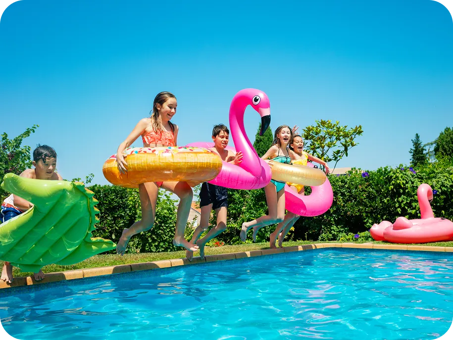Group of kids jumping into a backyard swimming pool with colorful inflatable toys on a sunny day.