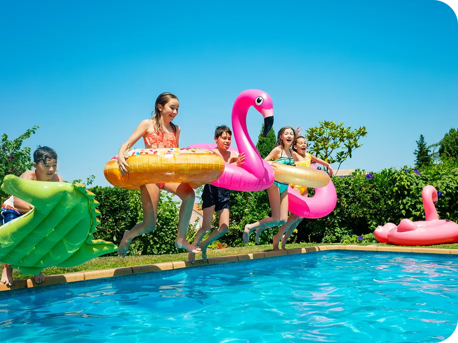 Group of kids jumping into a backyard swimming pool with colorful inflatable toys on a sunny day.