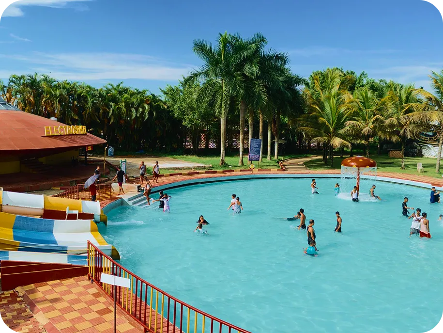 Families and children enjoying a waterpark pool with slides, palm trees, and interactive water features.