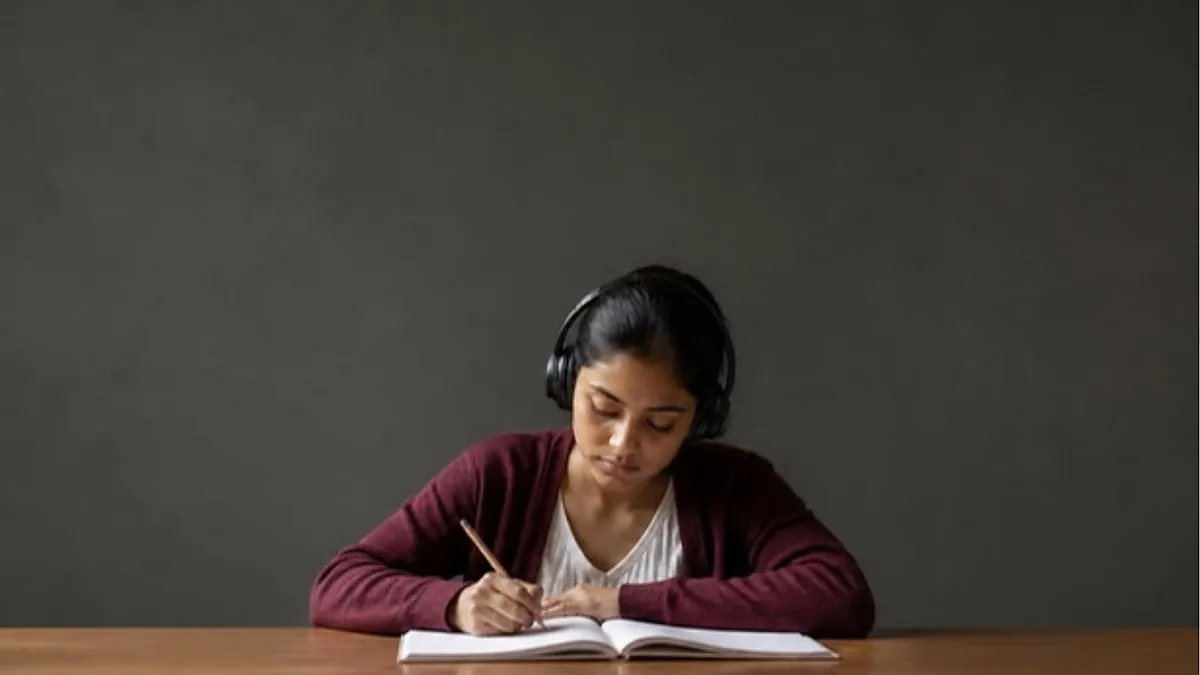 A student wearing headphones writes in an answer booklet at a desk in a quiet study room, practising for IELTS Academic vs General Training.