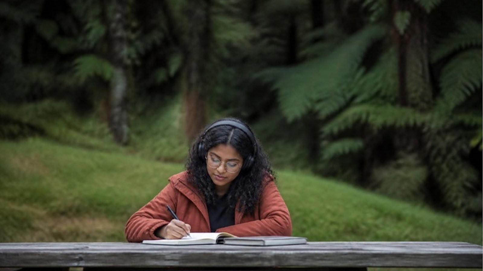 A student wearing headphones writes in a notebook at a bench among native bush on a New Zealand campus, preparing to meet New Zealand English language requirements.