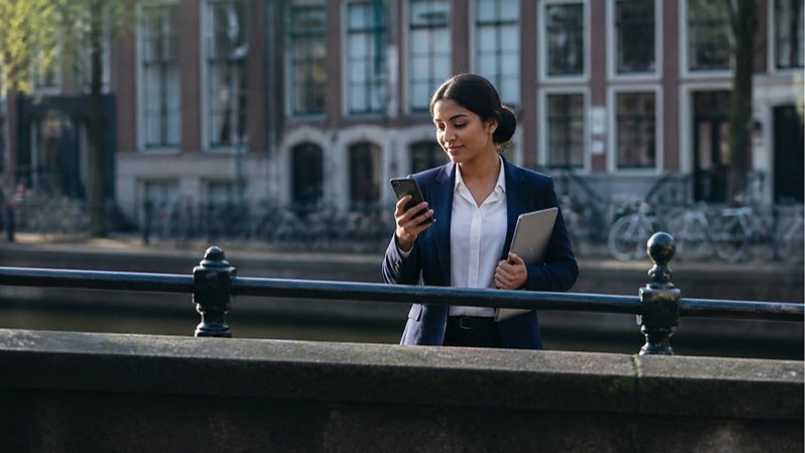 A graduate in a navy blazer checks her phone beside a Dutch canal, using her orientation year in the Netherlands to look for work.