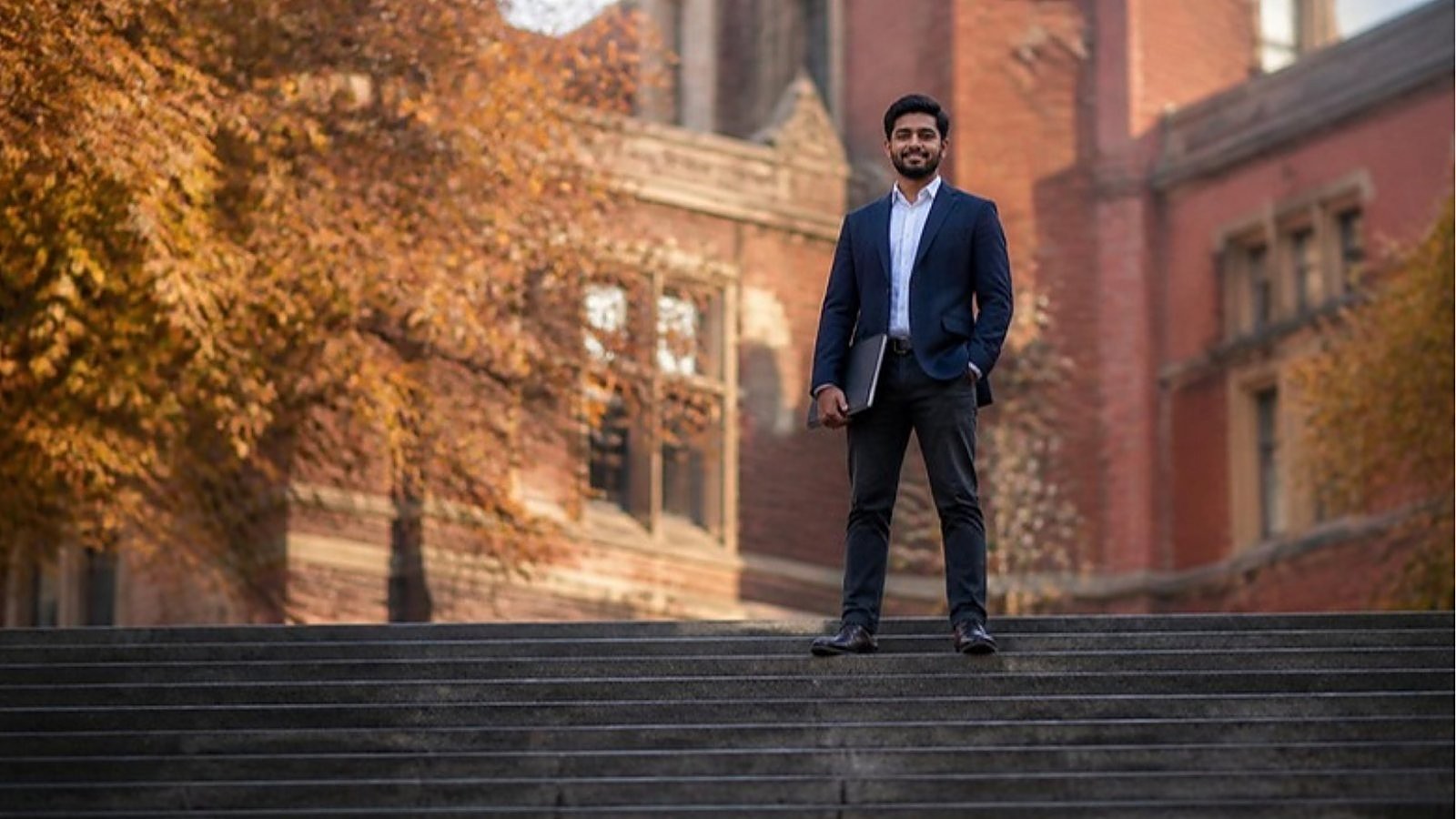A graduate in a navy blazer stands on stone steps in front of a red-brick UK university in autumn, ready to start a top-up degree in the UK.