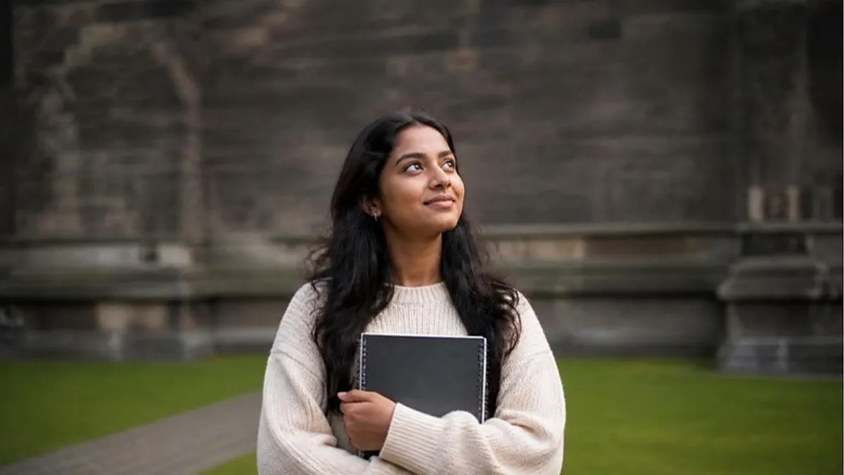 A student in a cream sweater holds a notebook and looks up hopefully on a campus lawn beside an old stone building, picturing her plan to study in Scotland.