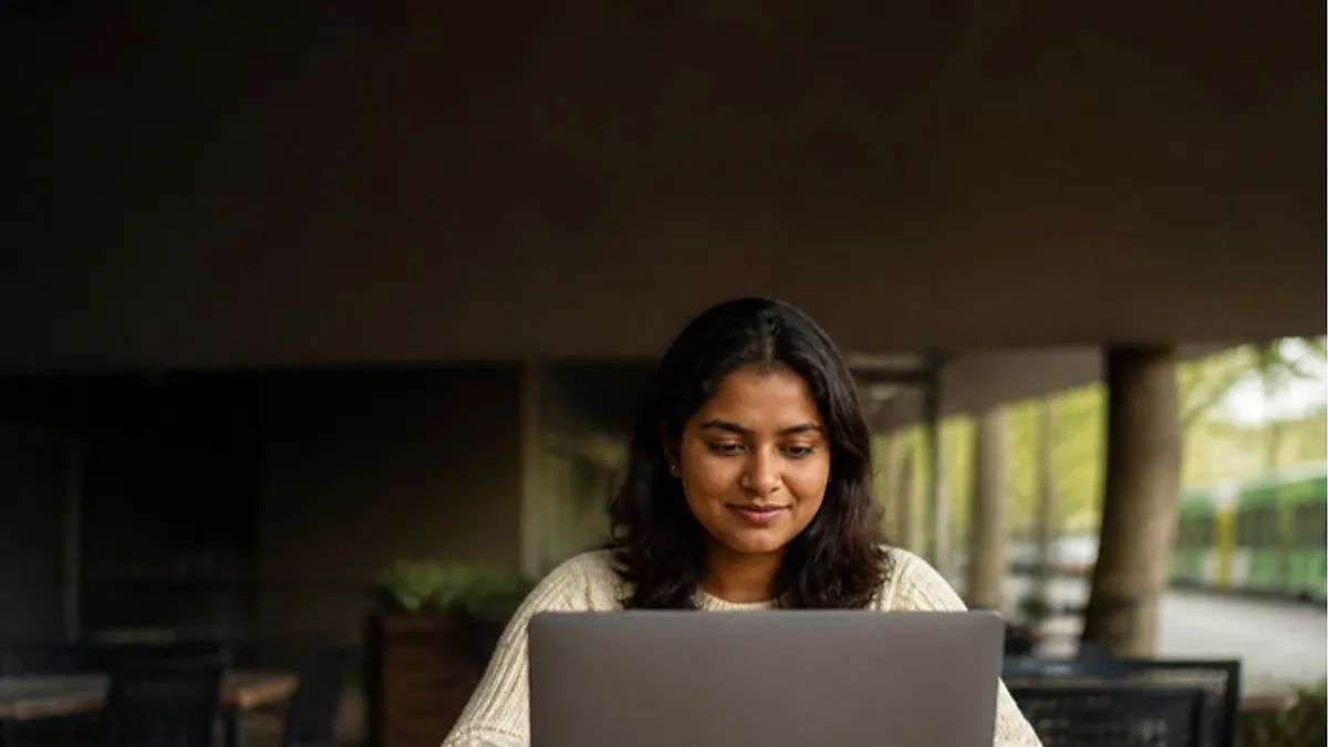 A young woman in a cream sweater works on a laptop at a Melbourne campus café with a tram passing behind, researching Swinburne University.