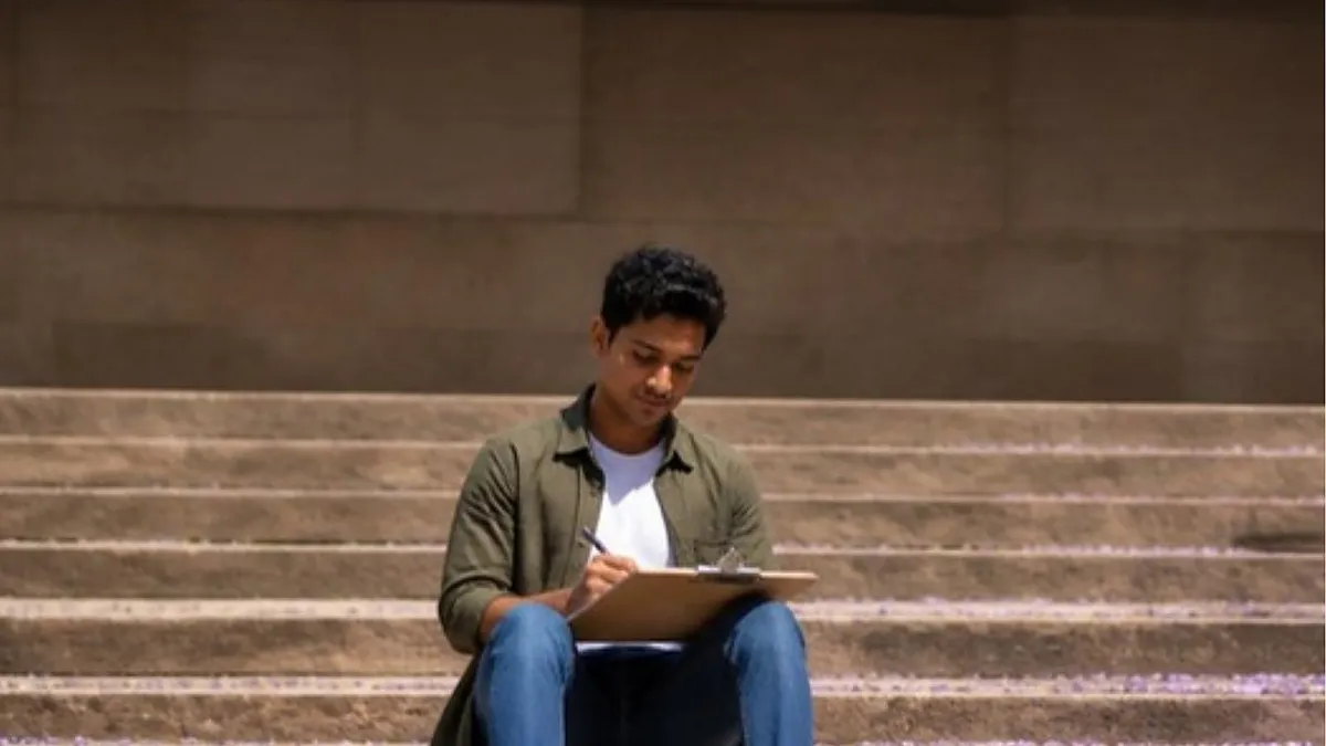 A student sits on sandstone steps on an Australian campus ticking items on a clipboard, working through his Australia student visa document checklist.