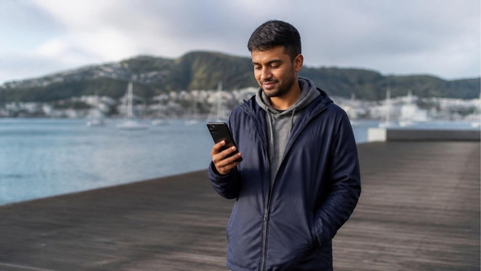 A student in a navy rain jacket reads his phone on the Wellington waterfront, checking what he needs to change education provider on a New Zealand student visa.
