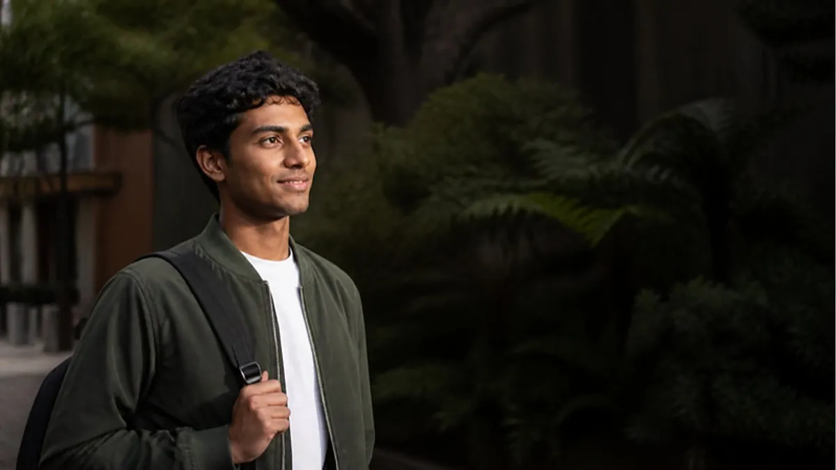A young man with a backpack stands among native ferns on a leafy Auckland campus, considering Auckland University of Technology.