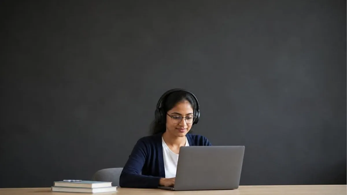 A student wearing headphones types on a laptop at a timber desk in a quiet study space while comparing PTE vs IELTS.