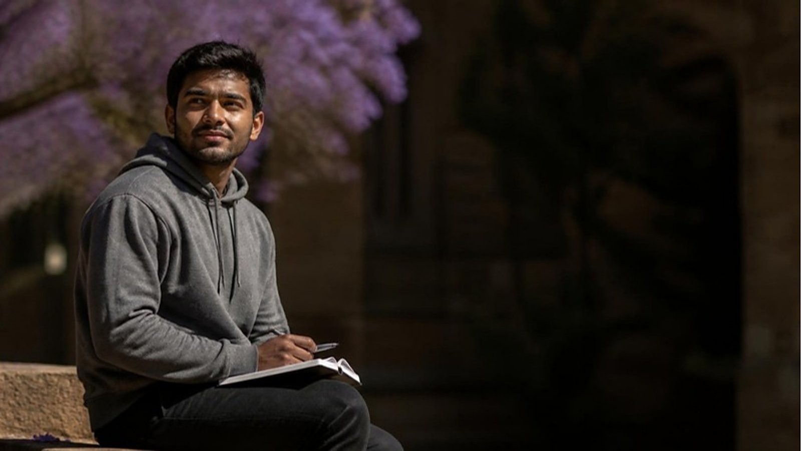 A student sits with a notebook under a jacaranda tree on an Australian university campus, planning his Australia student visa cost.
