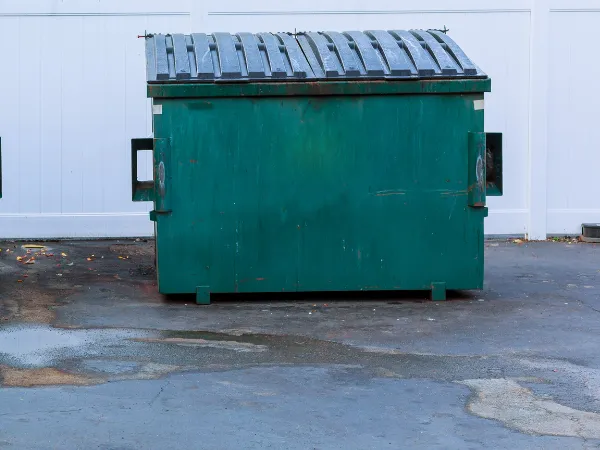 Empty green commercial dumpster placed on a paved surface against a white fence.