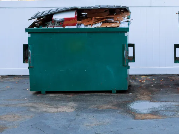 Overfilled commercial dumpster with cardboard and trash visible under an open lid.