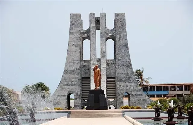 Bronze statue of a person stands before a tall, grey stone monument with arches