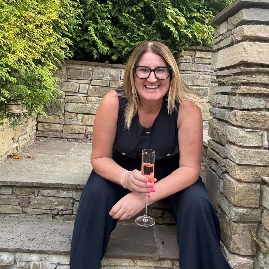 Emma Hine, a woman with blonde hair and glasses sitting at a table, smiling, in front of a brick wall