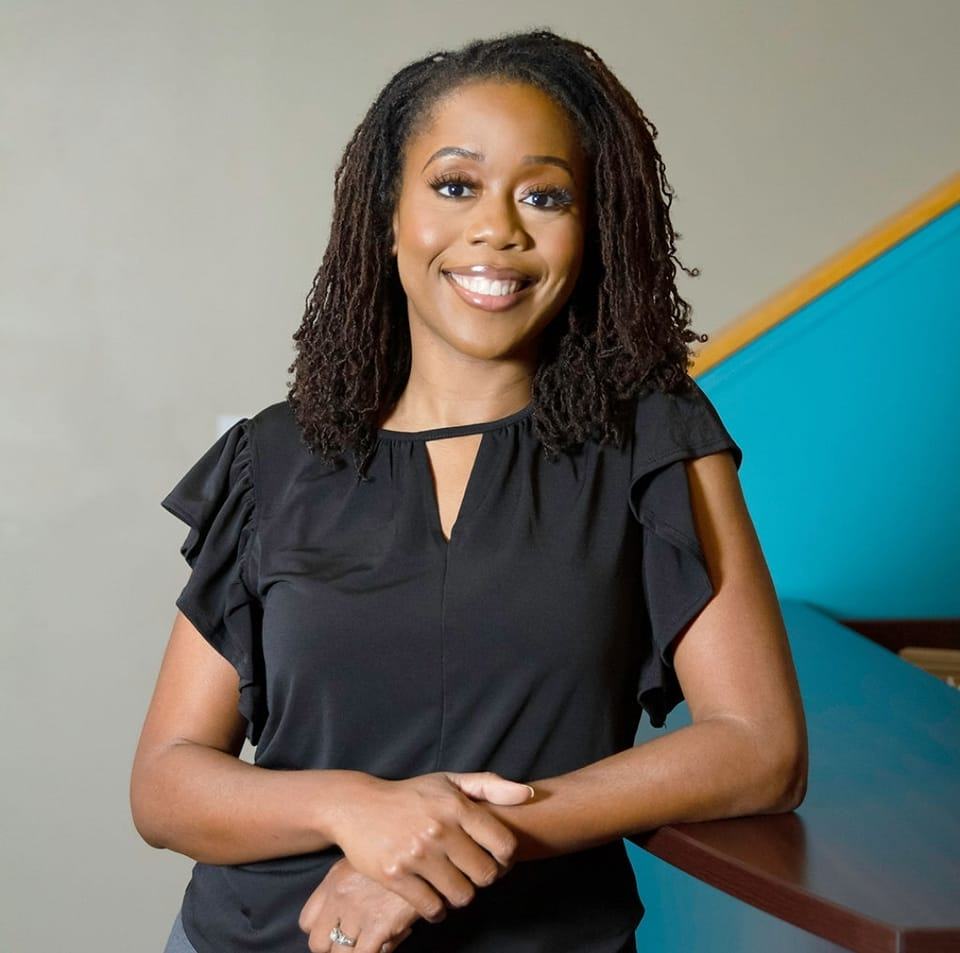 Portrait of Dr. Alicia James, a Black woman in her 40s with natural hair, wearing a vibrant patterned blazer, smiling warmly against a soft studio background. Her confident posture and direct gaze convey wisdom and approachability.