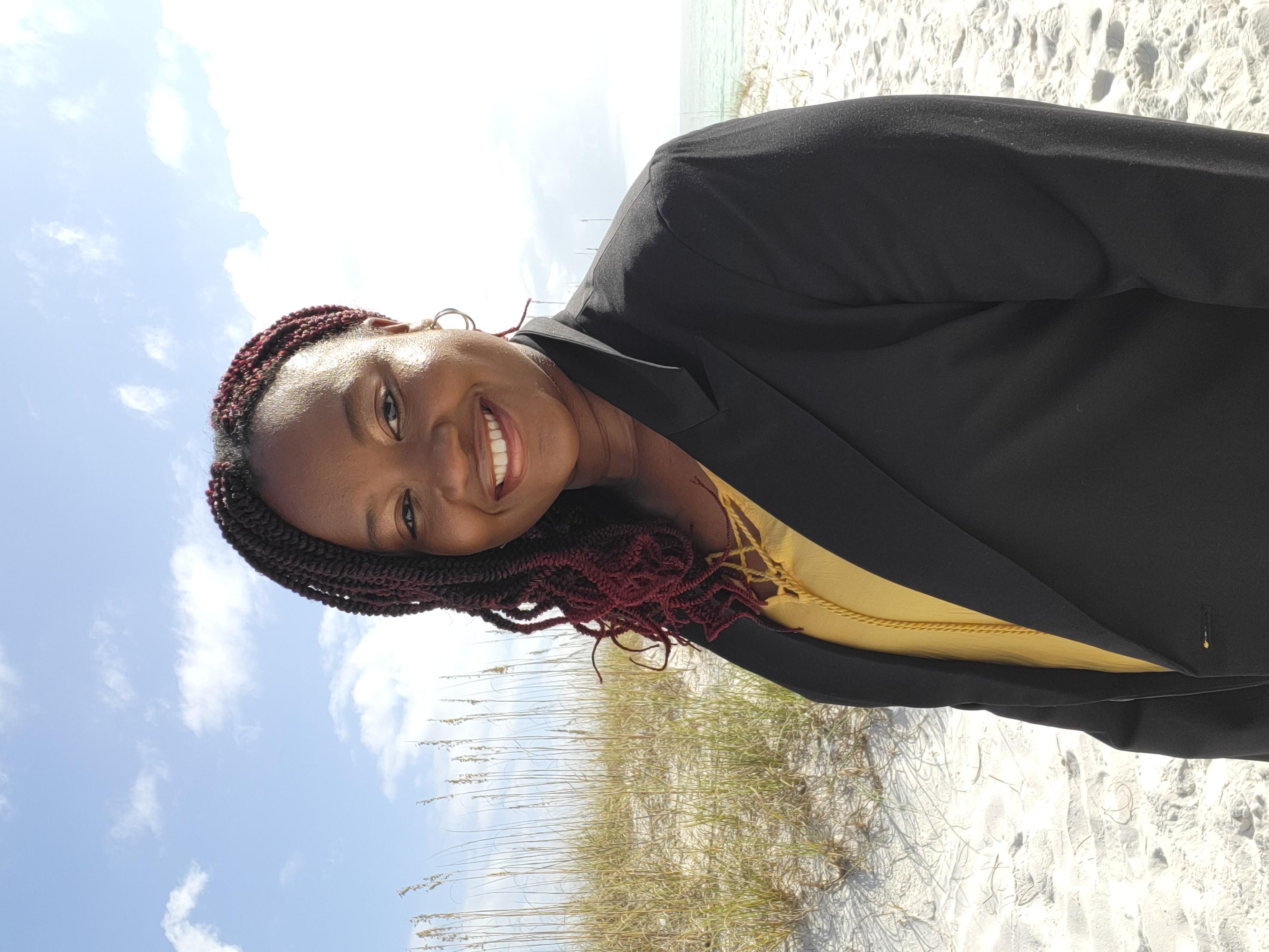 Portrait of Coach Maya Roberts, a Black woman in her 50s with silver-streaked locs, wearing a tailored black suit, seated in a modern chair. Her poised demeanor and gentle smile radiate wisdom and encouragement.