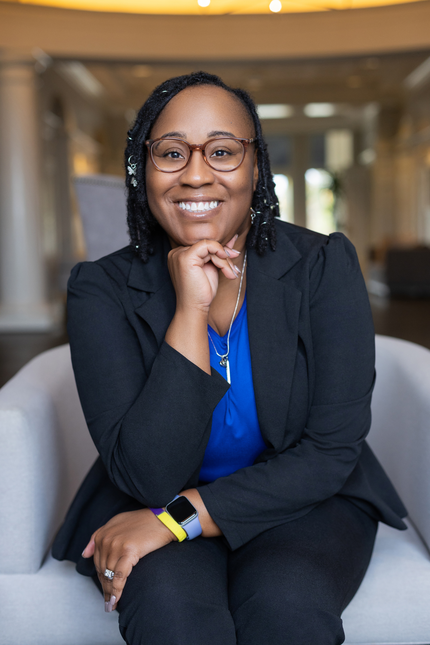 Portrait of Ms. Tiana Okafor, a young Black woman with short curly hair, wearing a sleek yellow dress, standing in front of a bookshelf. Her expression is thoughtful and inviting, suggesting expertise in personal development.
