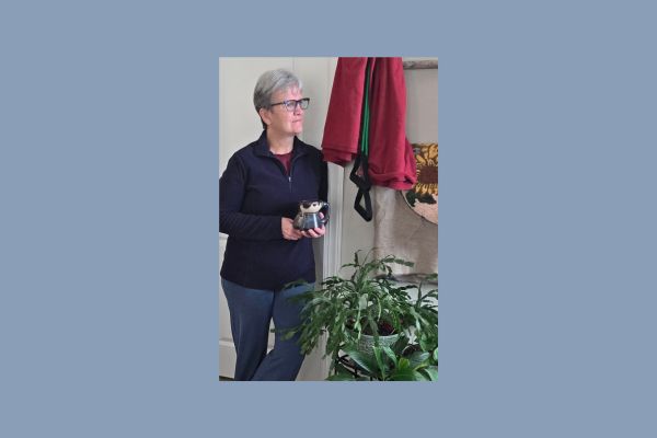 Claudette Eames standing calmly at home holding a mug beside houseplants, reflecting on nervous system regulation and rebuilding calm strength naturally.