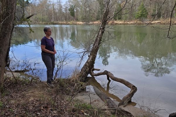 Claudette Eames standing quietly by a calm river surrounded by trees, reflecting in nature during a moment of peaceful solitude.