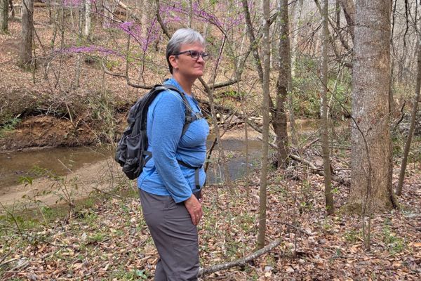 Woman hiking on a wooded trail with a backpack, pausing and listening to her body during recovery and rebuilding strength
