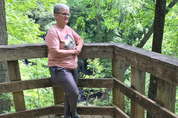 Claudette Eames standing on a wooden overlook surrounded by green forest, reflecting in a calm natural setting