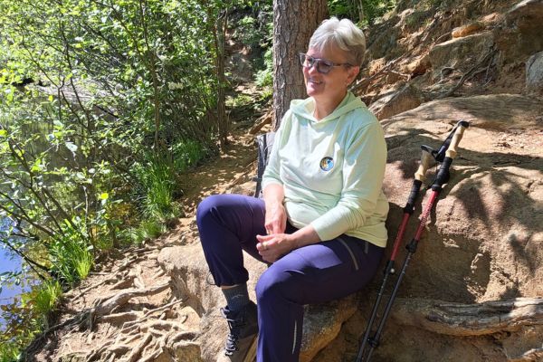 Claudette Eames sitting peacefully on a rock by a forest stream, smiling, surrounded by green trees and nature, holding hiking poles
