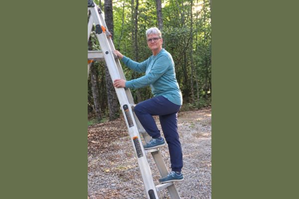 Claudette Eames standing on an aluminum ladder outdoors in a wooded setting, wearing a teal shirt and navy pants.
