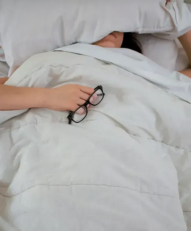  Woman hiding under a pillow in bed while holding her glasses, struggling with insomnia during menopause