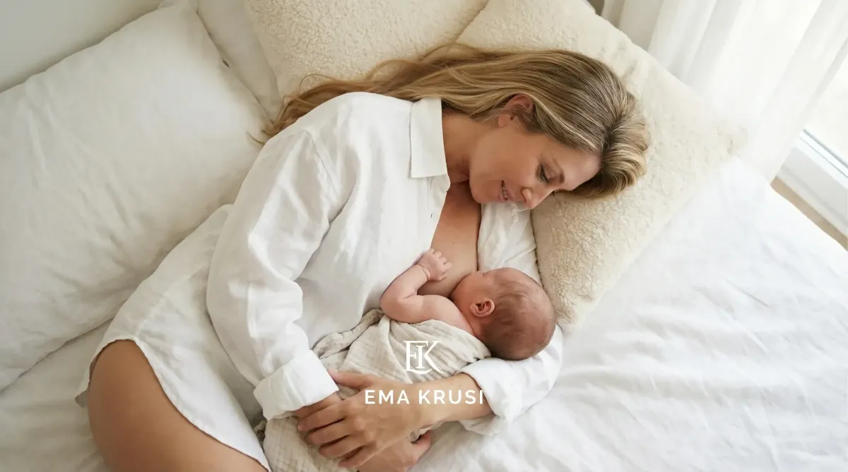 Mère allongée sur un lit allaitant son nouveau-né, souriant, dans une chambre blanche lumineuse.